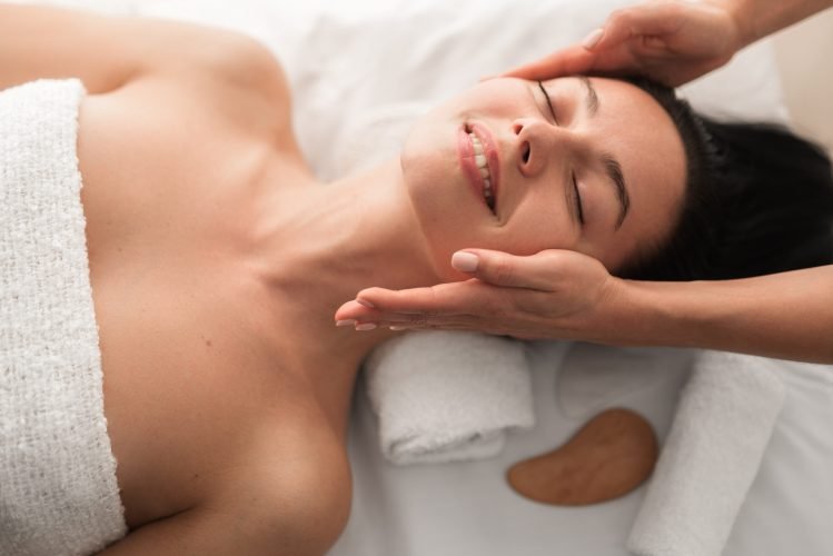 Top view of crop unrecognizable cosmetician doing face massage to cheerful young female customer with long dark hair, lying on couch with closed eyes and smiling